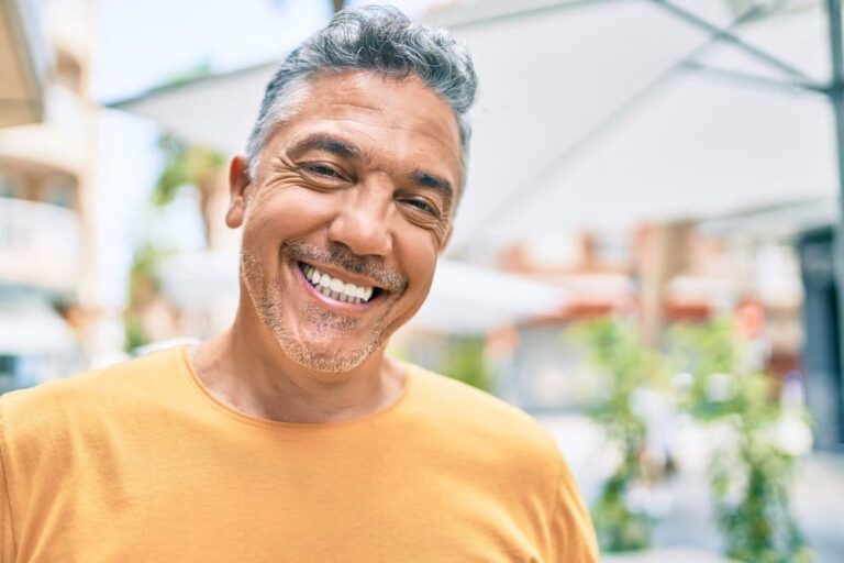 smiling man with gray hair and a yellow shirt standing in front of a building