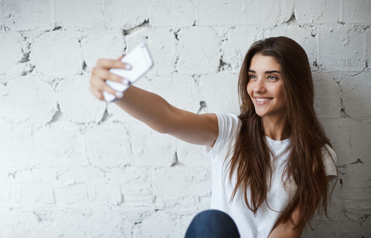 smiling woman holding up a white cell phone in front of a white brick wall