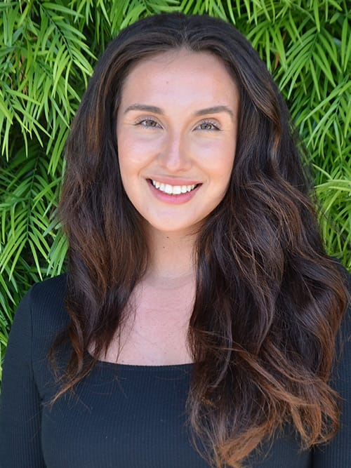 smiling woman with long dark hair and black top standing in front of a bush