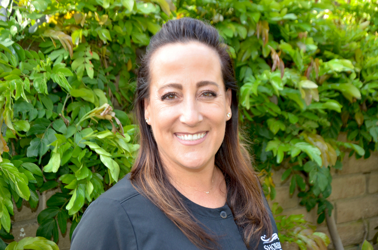 smiling woman with long brown hair and black shirt standing in front of a bush