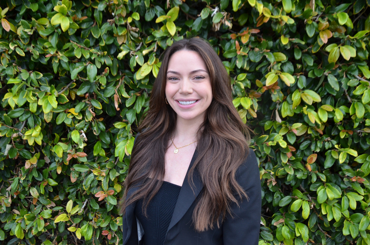 smiling woman in black jacket and black dress standing in front of a hedge