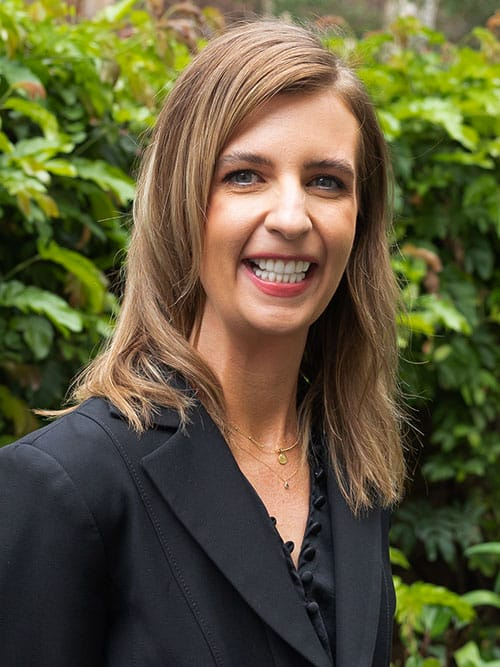 smiling woman in black jacket and black blouse standing in front of bushes