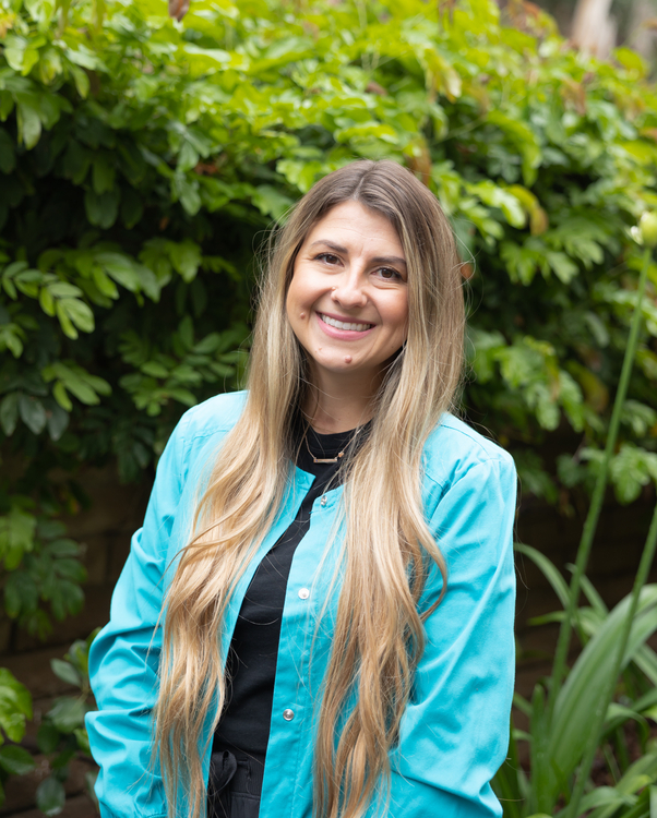 smiling woman in blue jacket and black tie standing in front of bushes