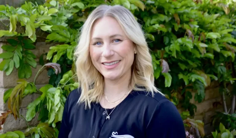 blond woman with blue eyes and a black shirt standing in front of a bush