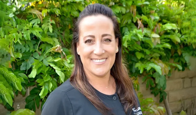 smiling woman with long brown hair and black shirt standing in front of a bush