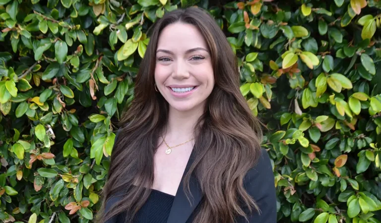 smiling woman in black jacket and black top standing in front of a bush