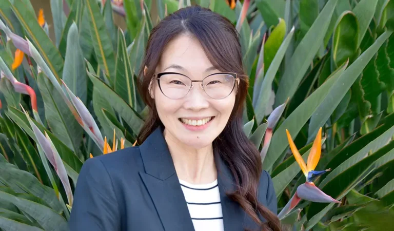 smiling woman in glasses standing in front of a garden of flowers