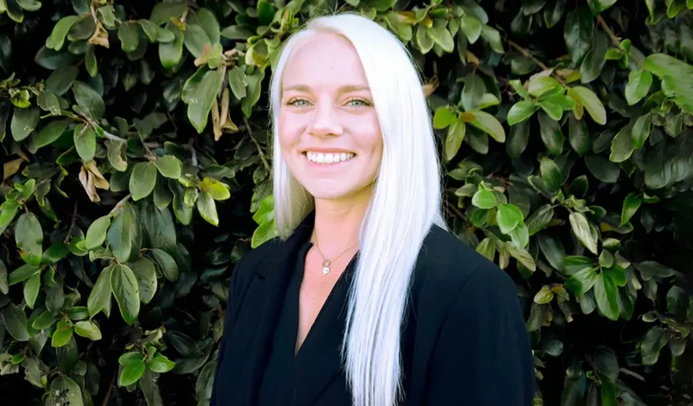 blond woman with white hair and black shirt standing in front of a bush