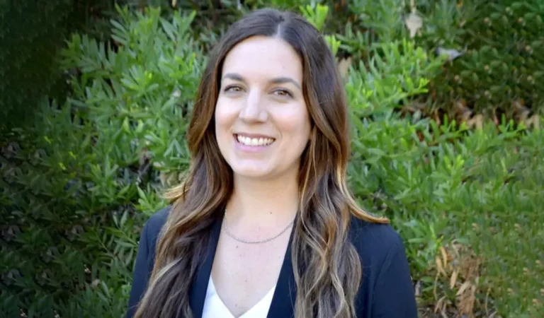 smiling woman in black jacket and white shirt standing in front of bushes