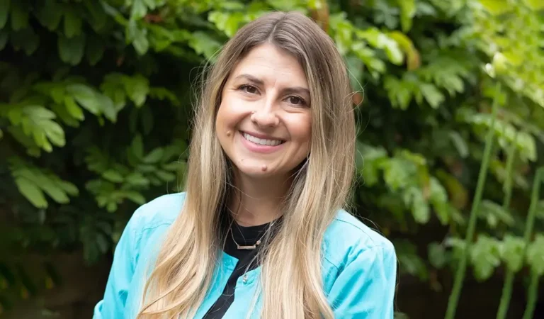 smiling woman with long blonde hair and blue shirt standing in front of a bush