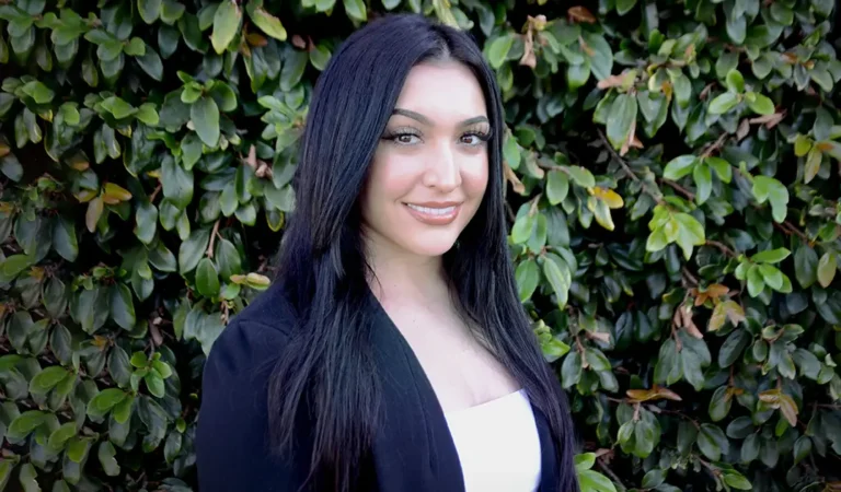 smiling woman with long black hair and black blazer standing in front of a bush