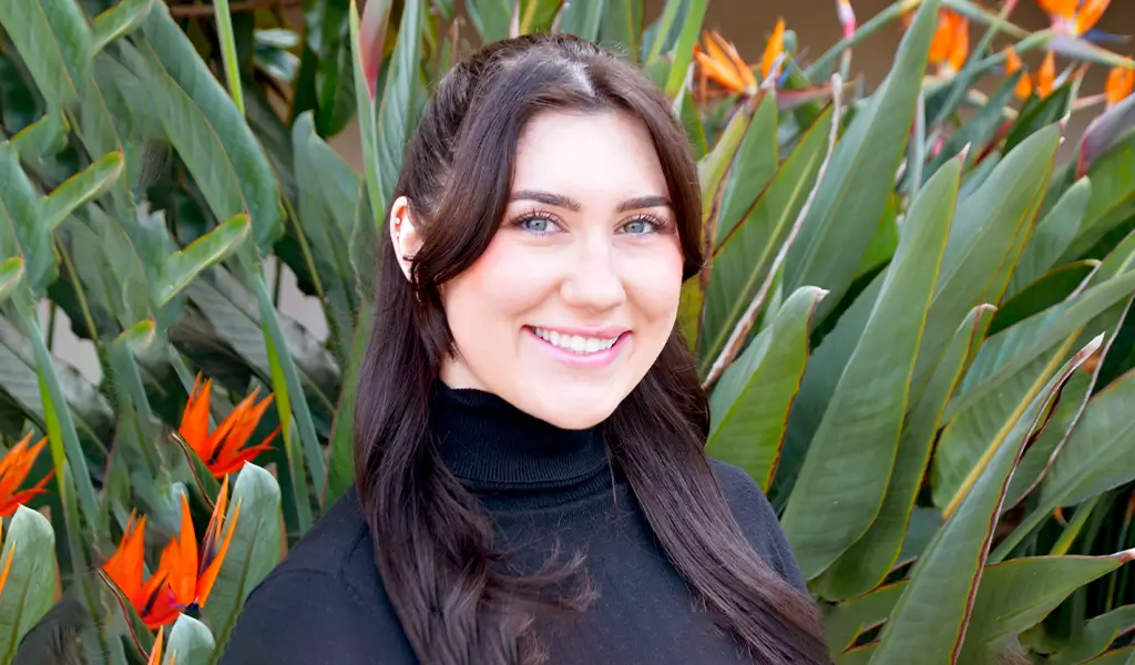 smiling woman in black turtle neck top standing in front of a bush of flowers