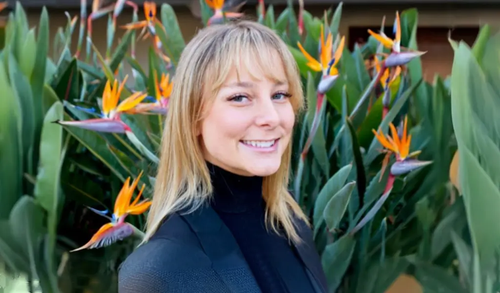 smiling woman in black jacket standing in front of a bunch of flowers