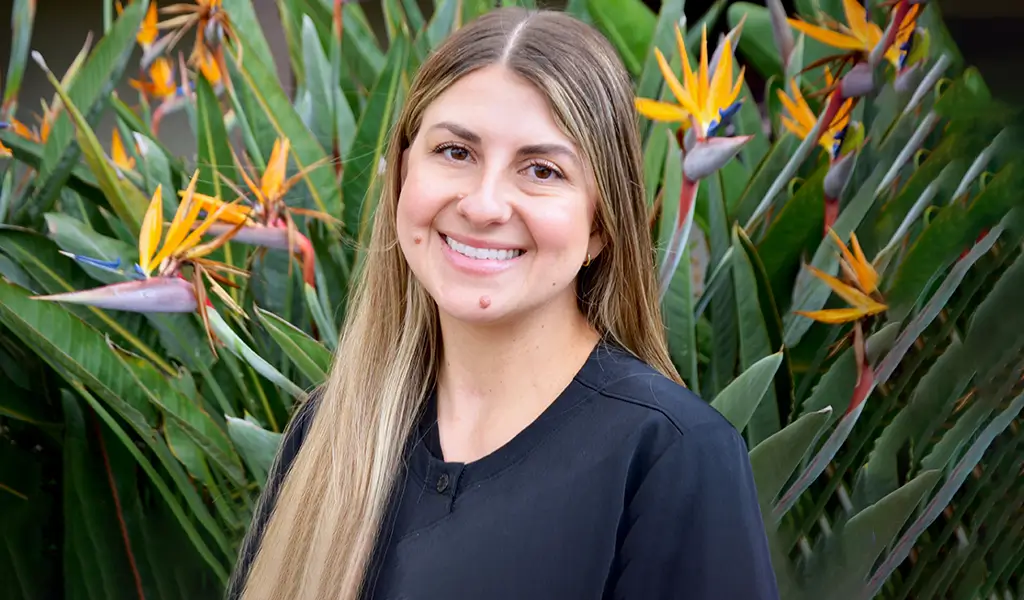 smiling woman with long blonde hair standing in front of a flower garden